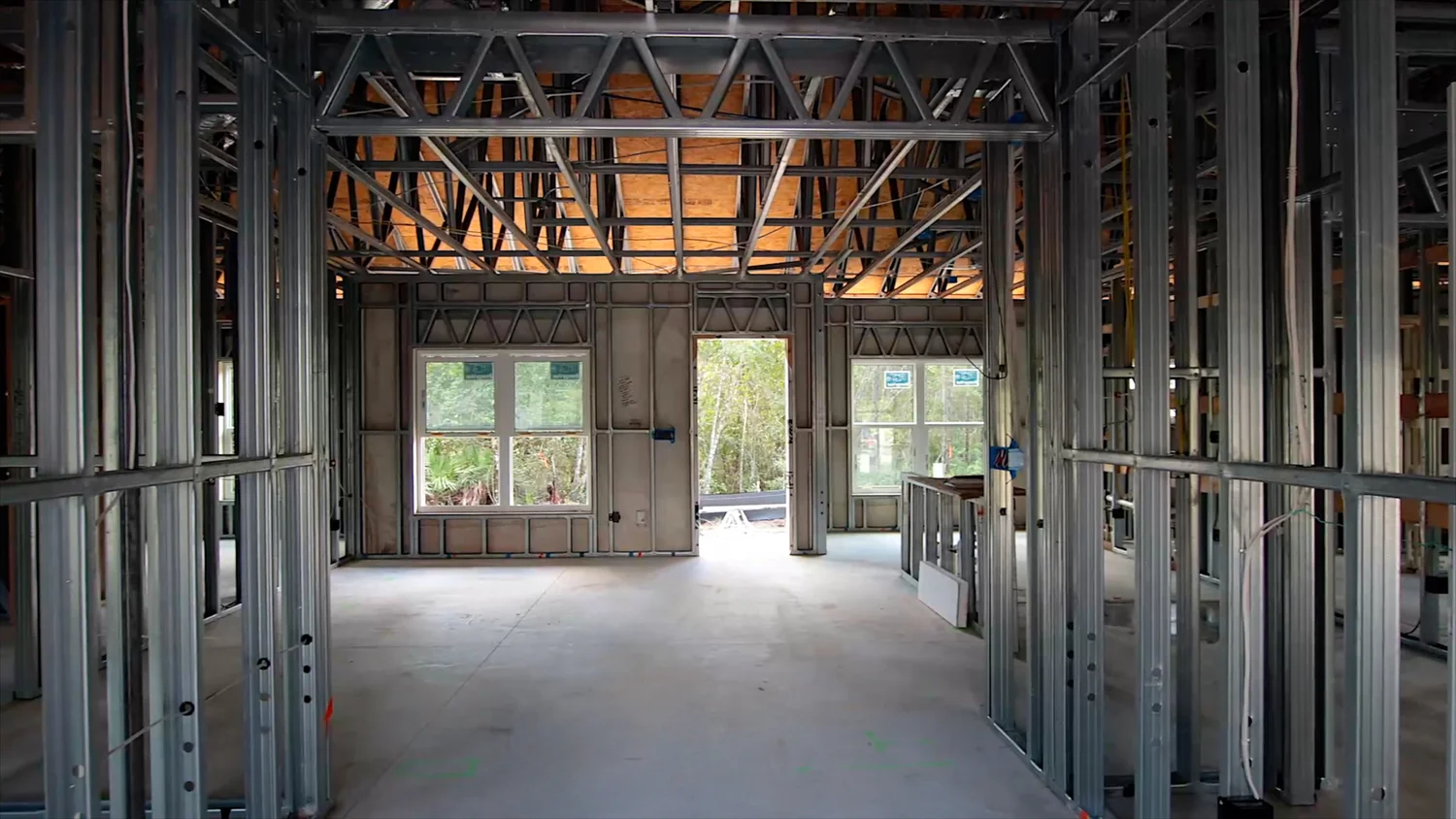 Interior view of a steel frame home under construction showing open floor plan, exposed ceiling trusses with wood sheathing above, and concrete slab floor